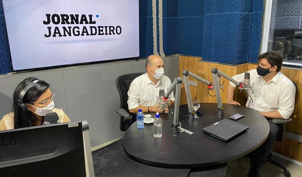 prefeito usando máscara, sentado entre um rapaz e uma moça também de máscara, em estúdio de rádio